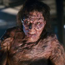 Close-up of a grotesque, scarred humanoid creature with wet hair and mottled skin in a dim industrial setting.