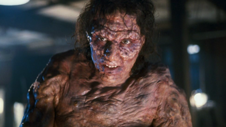 Close-up of a grotesque, scarred humanoid creature with wet hair and mottled skin in a dim industrial setting.