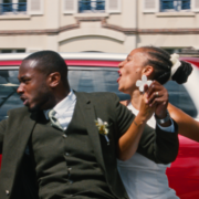A sharply dressed man and a woman dance exuberantly beside a red car on a city street, their raised arms and lively poses conveying joy.