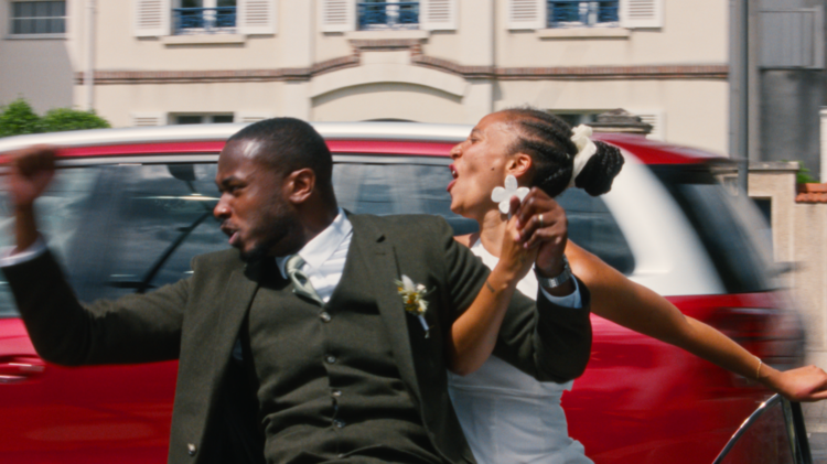 A sharply dressed man and a woman dance exuberantly beside a red car on a city street, their raised arms and lively poses conveying joy.