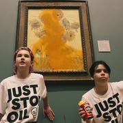 Two people in “JUST STOP OIL” shirts pose before a vandalized sunflower painting in a museum; one holds a can of soup.