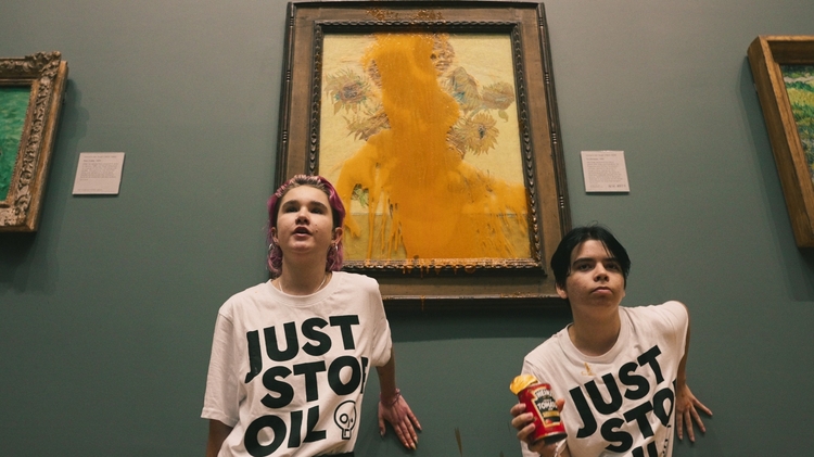 Two people in “JUST STOP OIL” shirts pose before a vandalized sunflower painting in a museum; one holds a can of soup.