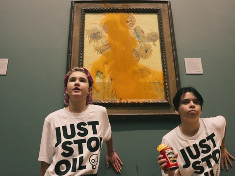 Two people in “JUST STOP OIL” shirts pose before a vandalized sunflower painting in a museum; one holds a can of soup.