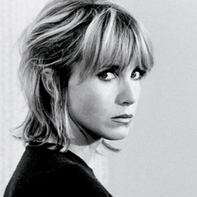 Black-and-white portrait of a blonde woman with bangs, looking over her shoulder against a plain background.