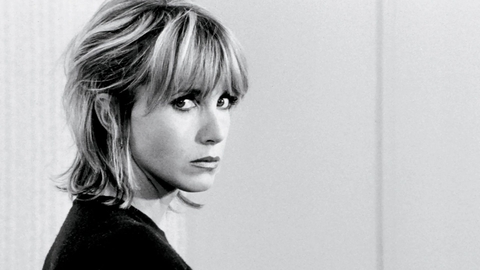Black-and-white portrait of a blonde woman with bangs, looking over her shoulder against a plain background.