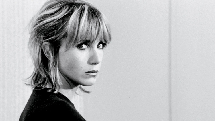 Black-and-white portrait of a blonde woman with bangs, looking over her shoulder against a plain background.