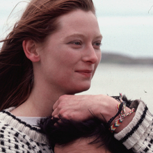 Close-up of a smiling woman with windblown hair hugging someone on a beach.