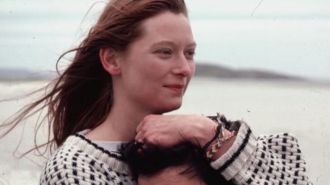 Close-up of a smiling woman with windblown hair hugging someone on a beach.