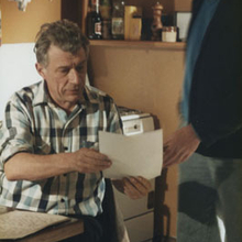 An older man in a plaid shirt sits at a table reading a document as a younger man stands beside him.