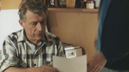 An older man in a plaid shirt sits at a table reading a document as a younger man stands beside him.