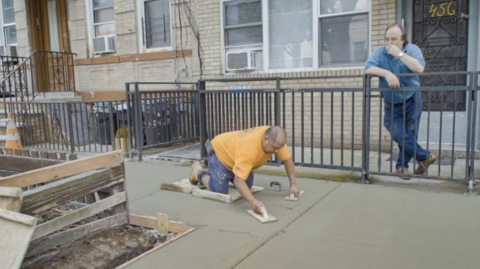 Construction worker kneels while smoothing a freshly poured sidewalk in front of a brick townhouse; another worker watches nearby.