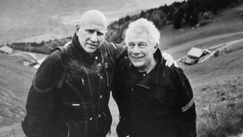 A black and white photo of two men standing outside with their arms around each other.