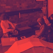 In a red-tinted workshop, several people crouch around large sheets on the floor, focused on a project beside equipment and circular artwork.