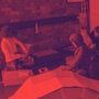 In a red-tinted workshop, several people crouch around large sheets on the floor, focused on a project beside equipment and circular artwork.