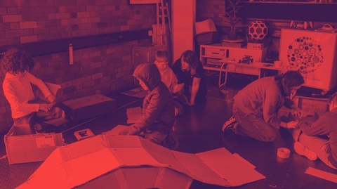 In a red-tinted workshop, several people crouch around large sheets on the floor, focused on a project beside equipment and circular artwork.