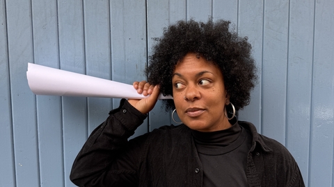 A photo of a woman holding a paper cone up to her ear as if she is listening to it.