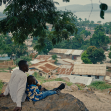 Teenager and child sit beneath a tree on a rocky hillside, overlooking a lush lakeside village with mountains beyond.