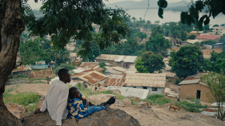 Teenager and child sit beneath a tree on a rocky hillside, overlooking a lush lakeside village with mountains beyond.