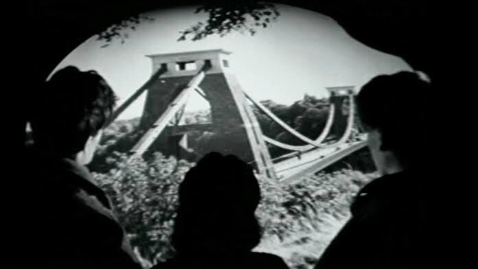 Black-and-white view of three silhouetted people watching footage of a suspension bridge projected on screen.