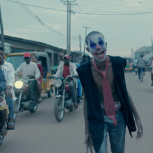 A boy standing in the middle of a road with clown makeup on as motorbikes go past.