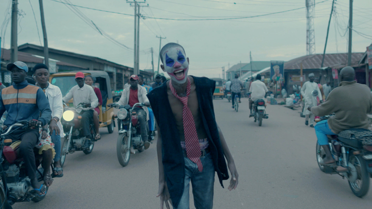 A boy standing in the middle of a road with clown makeup on as motorbikes go past.