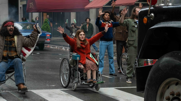 A lady in a wheelchair holds her hands up in defiance with a group of other people in front of a truck.