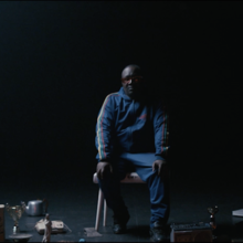 Man in a blue tracksuit sits on a chair in a dim stage setting, surrounded by scattered props and lighting equipment.