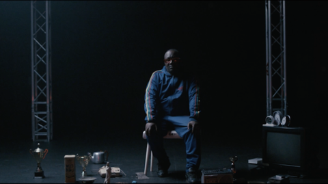 Man in a blue tracksuit sits on a chair in a dim stage setting, surrounded by scattered props and lighting equipment.