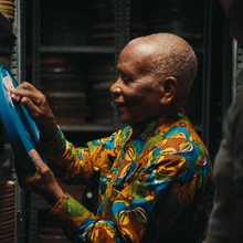 Man in colourful patterned clothing reaches toward a blue oxygen tank beside stacked black barrels.