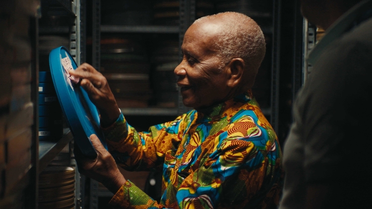 Man in colourful patterned clothing reaches toward a blue oxygen tank beside stacked black barrels.