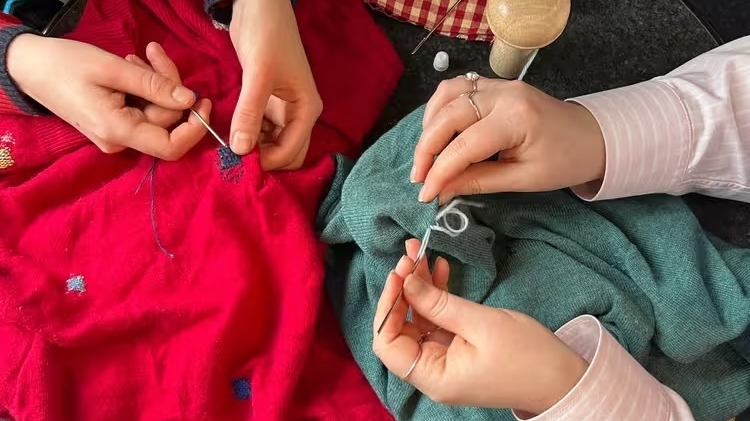 Two people embroider colourful sweaters by hand, surrounded by yarn, needles, and a pincushion.