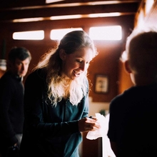 Smiling woman with a ponytail hands a napkin to a silhouetted person in a warmly lit room.