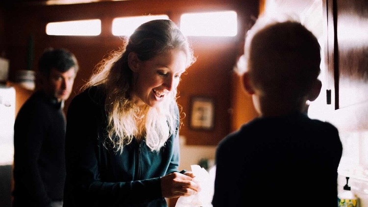 Smiling woman with a ponytail hands a napkin to a silhouetted person in a warmly lit room.