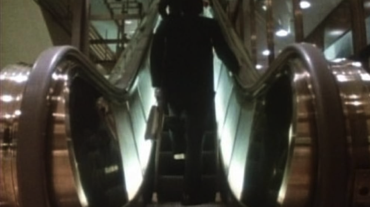 Silhouetted passengers ride an escalator upward inside a brightly lit modern building.