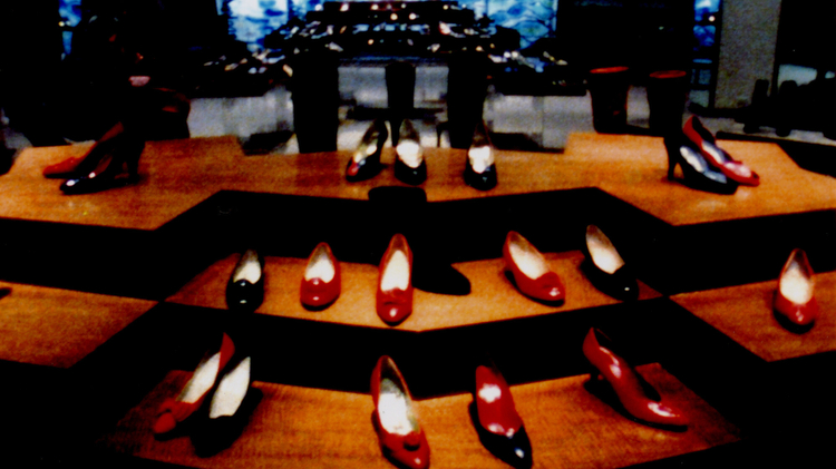 Rows of red, black, and white high-heeled shoes displayed on wooden platforms in a dimly lit setting.