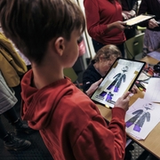 A child in a red hoodie views a fashion sketch on a tablet at a busy art workshop, surrounded by paper designs and colourful drawing tools.