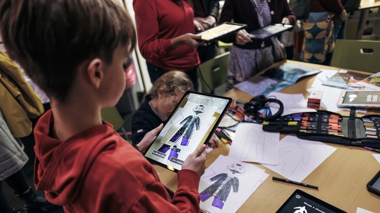 A child in a red hoodie views a fashion sketch on a tablet at a busy art workshop, surrounded by paper designs and colourful drawing tools.