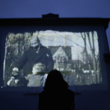 Silhouetted viewer watches a black-and-white film projected on a large screen in a dark theatre.