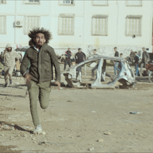 A man runs toward the camera through a debris-strewn street, with a wrecked car and onlookers in front of graffitied buildings.