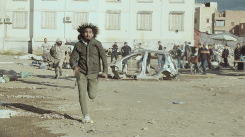 A man runs toward the camera through a debris-strewn street, with a wrecked car and onlookers in front of graffitied buildings.