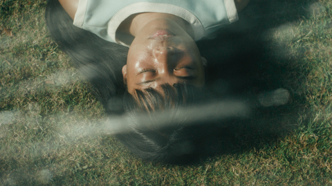 Person with eyes closed lying on grass, partially covered by a light-coloured garment and long dark hair.