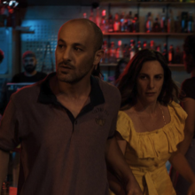A bald man and woman in a yellow dress walk through a dimly lit bar with colourful red and blue lighting.