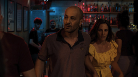 A bald man and woman in a yellow dress walk through a dimly lit bar with colourful red and blue lighting.