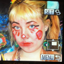 An image of the Raina Griefer a white artist with blonde hair and clown makeup on rendered in a 90's VHS style.