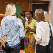 A Black woman with a soft smile and french plaits smiles softly in a conversation with two white women with blonde hair who have their backs to the viewer.