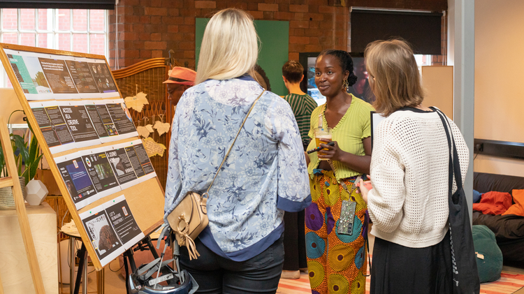 A Black woman with a soft smile and french plaits smiles softly in a conversation with two white women with blonde hair who have their backs to the viewer.