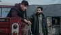 Barry Keoghan looks off into the distance sitting in the back of a red van, whilst Christopher Abott looks in his direction whilst leaning on the red van.