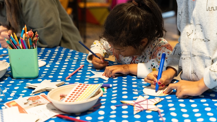 A young girl colours in a star with a blue pencil ontop of a spotty blue table cloth.