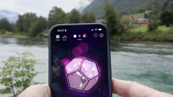 A beautiful landscape on the river with a person holding up a mobile screen of Games, Synths and Impossible Instruments.