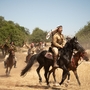 Horsemen in traditional attire traverse a dusty field, wielding rifles and surrounded by lush trees under a clear blue sky.